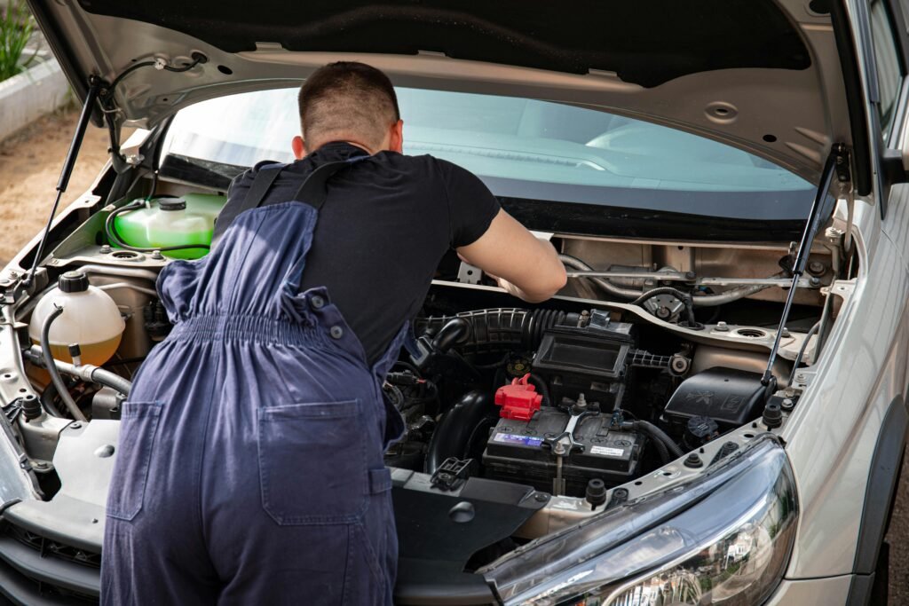 Mechanic in overalls working on a car engine outdoors, showcasing vehicle repair skills.